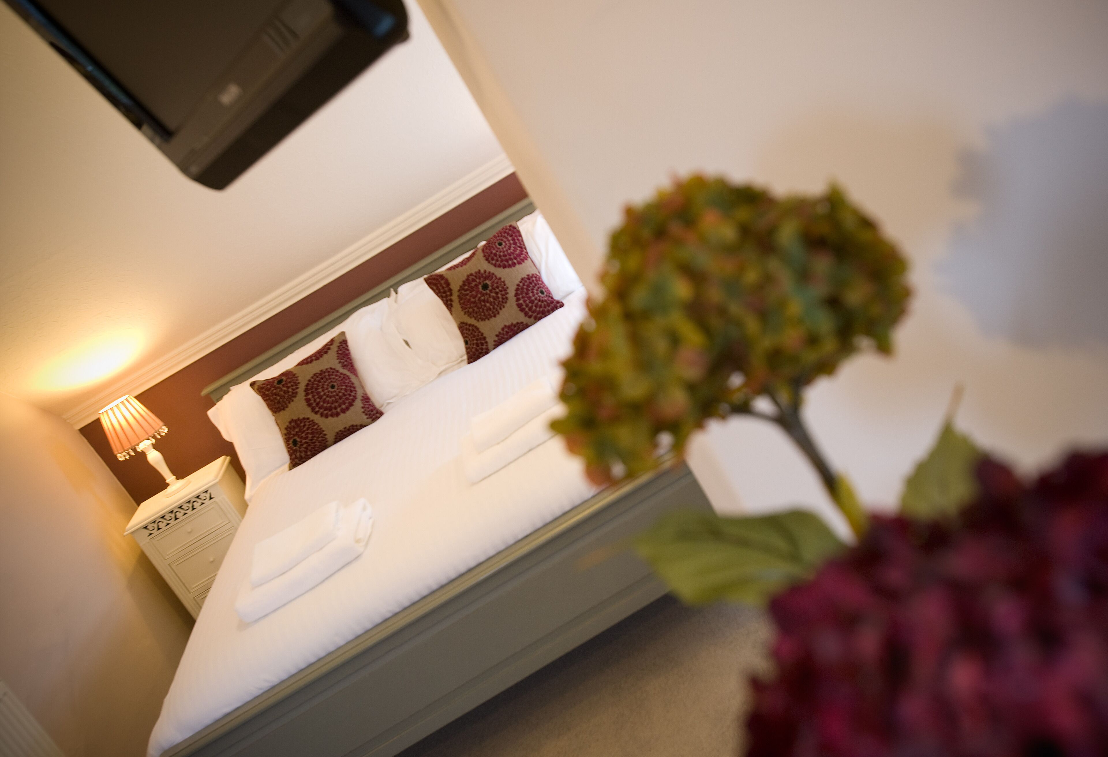 A neatly made bed with white linens and decorative pillows, a bedside lamp, and a small potted plant.