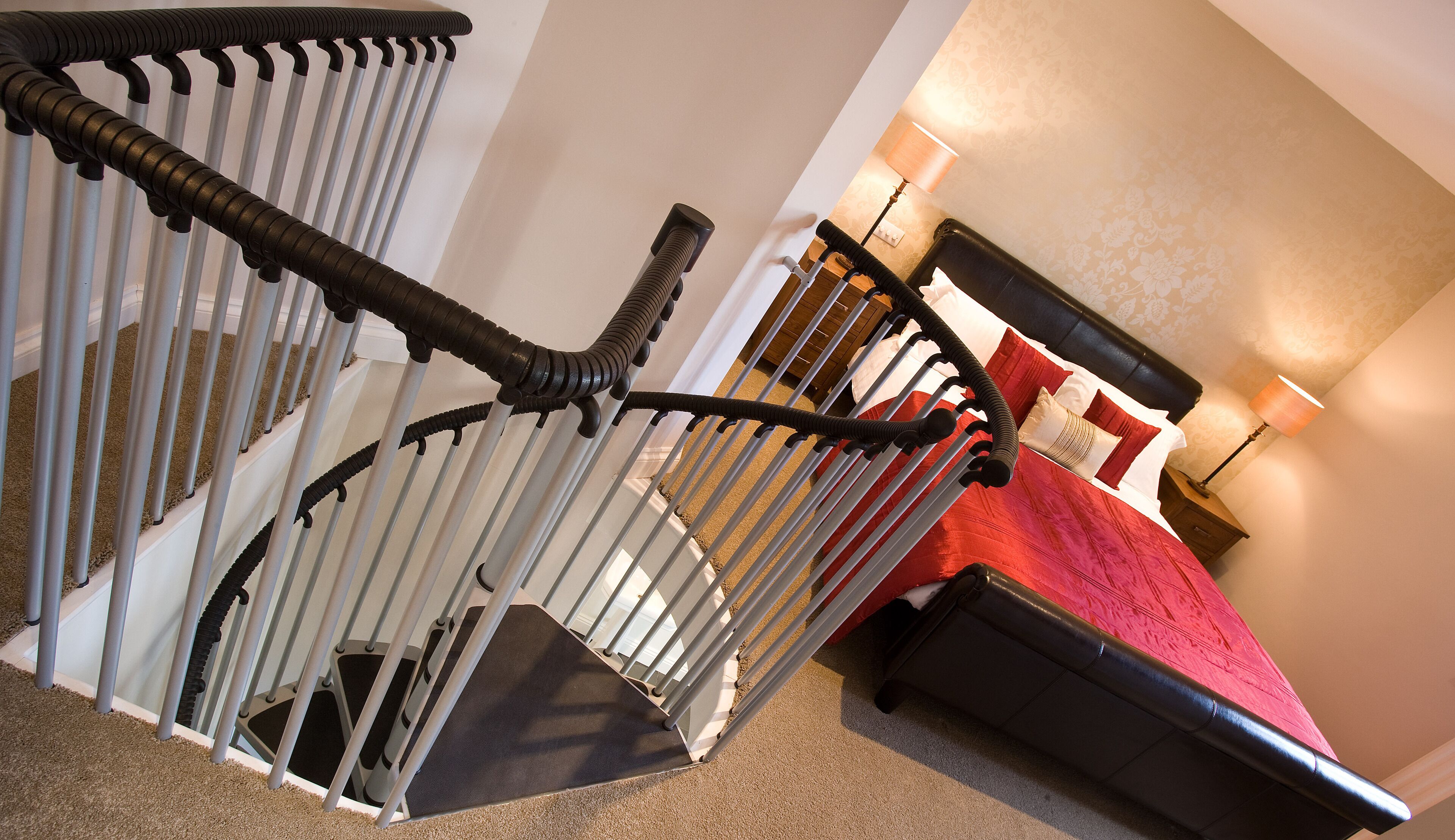 Un escalier avec une rampe, des marches recouvertes de moquette, et un lit avec une literie rouge dans la pièce adjacente.