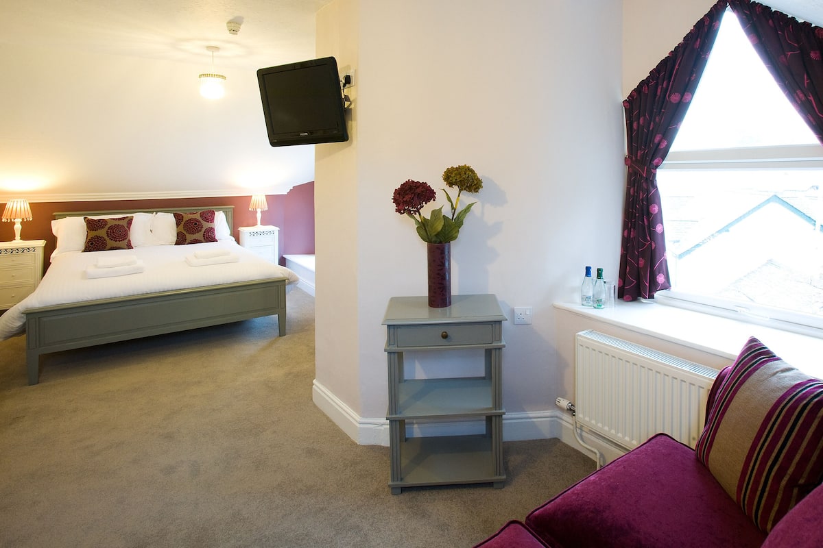 Une chambre d’hôtel avec un lit, une table de chevet, une petite table sur laquelle est posé un vase de fleurs, une fenêtre avec des rideaux et un radiateur.