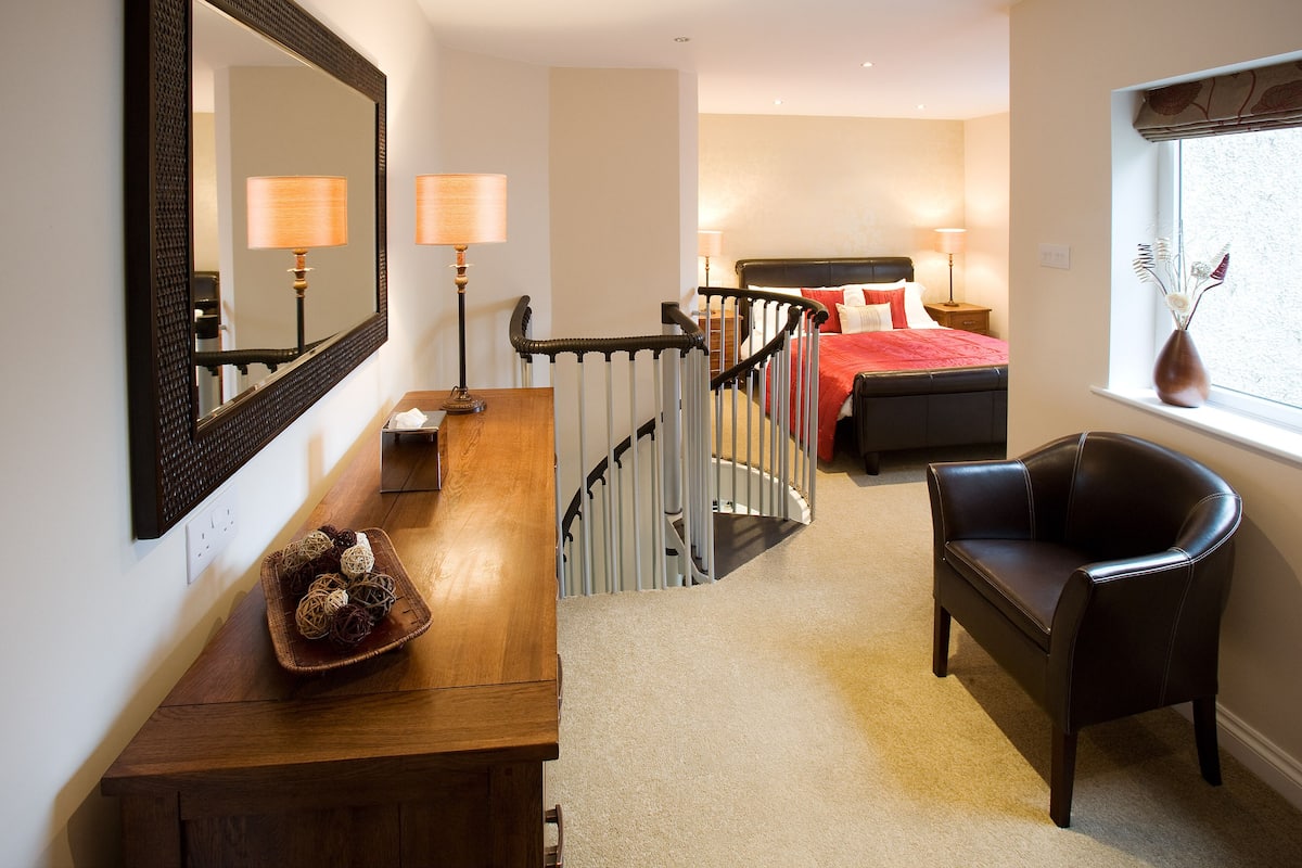 Une chambre d’hôtel avec un bureau en bois, une chaise, un lit recouvert d’une courtepointe rouge, une table de chevet avec des lampes, une fenêtre donnant sur un paysage agréable et un escalier.