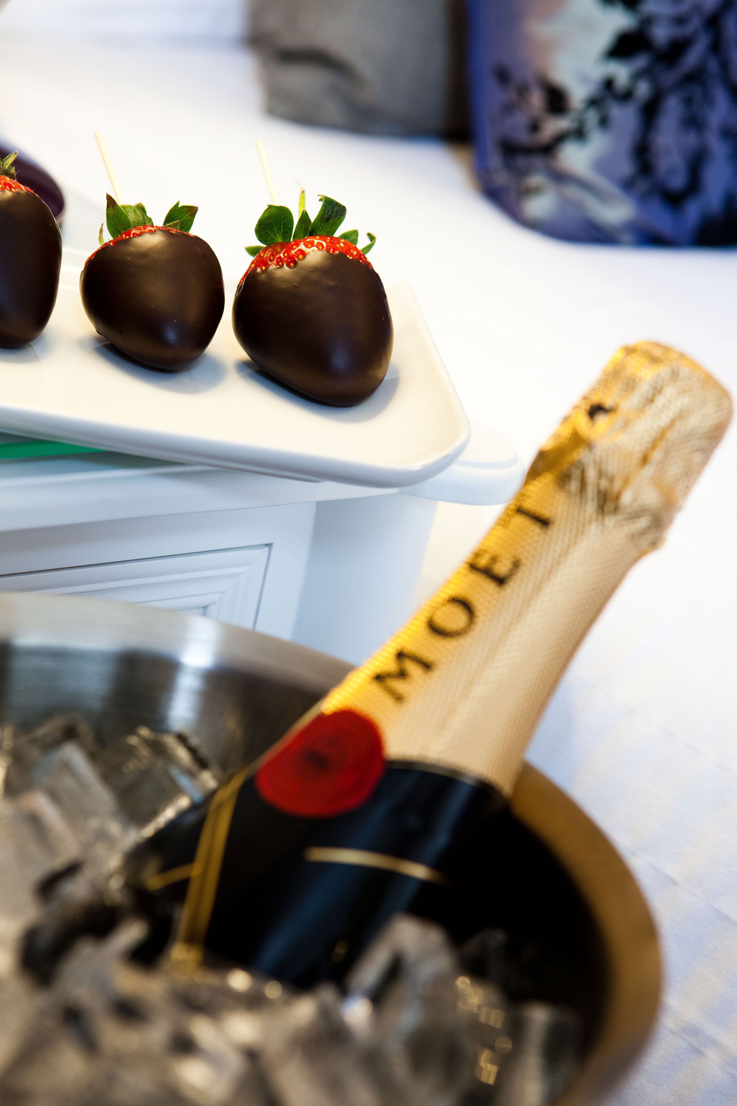 A bottle of champagne with a label, ice cubes, and three chocolate-covered strawberries with skewers.