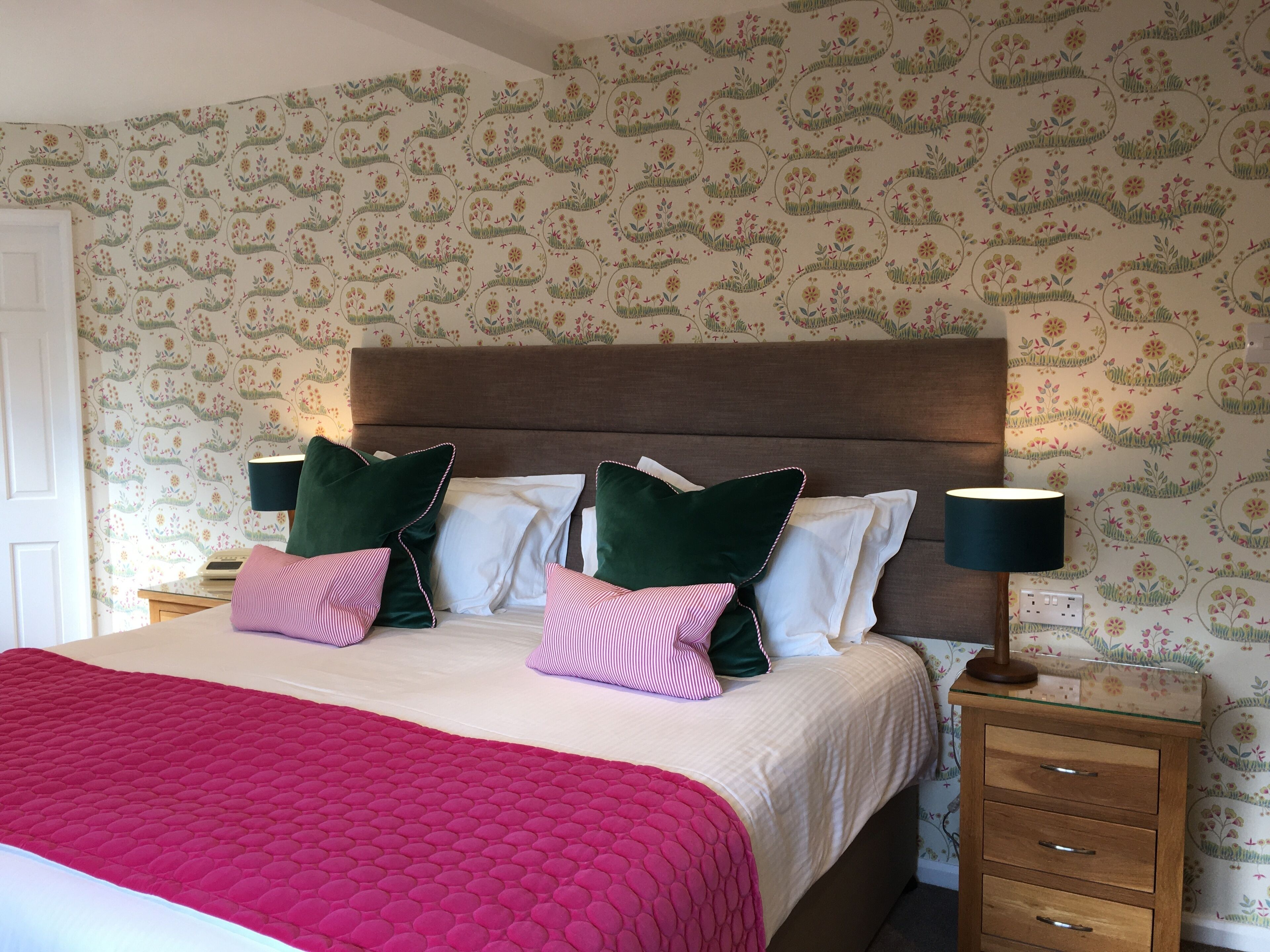A neatly made bed with a pink quilt, green and white pillows, and a wooden nightstand with a lamp.