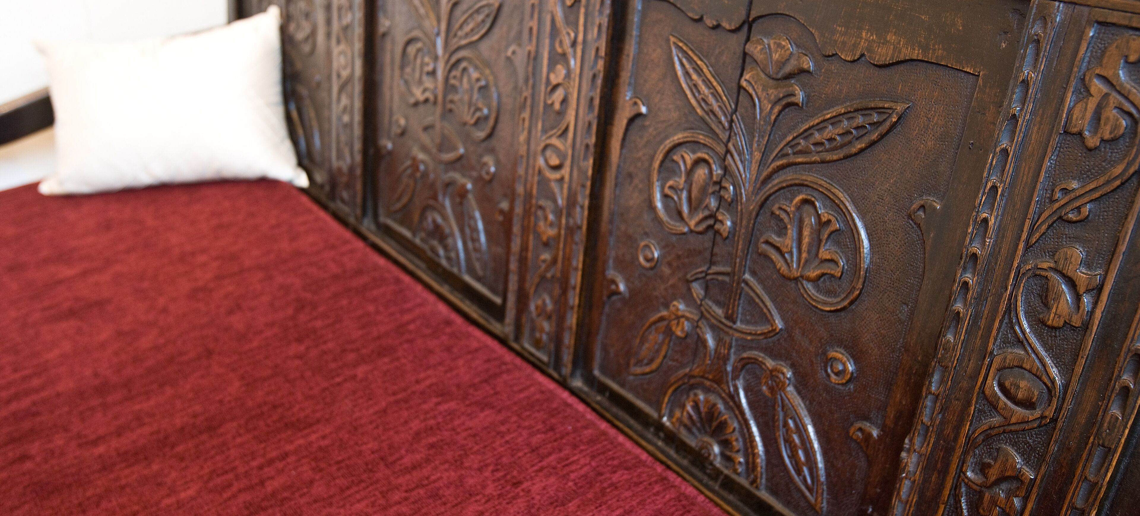 A bed with a red cover and a white pillow, a wooden headboard with floral carvings.