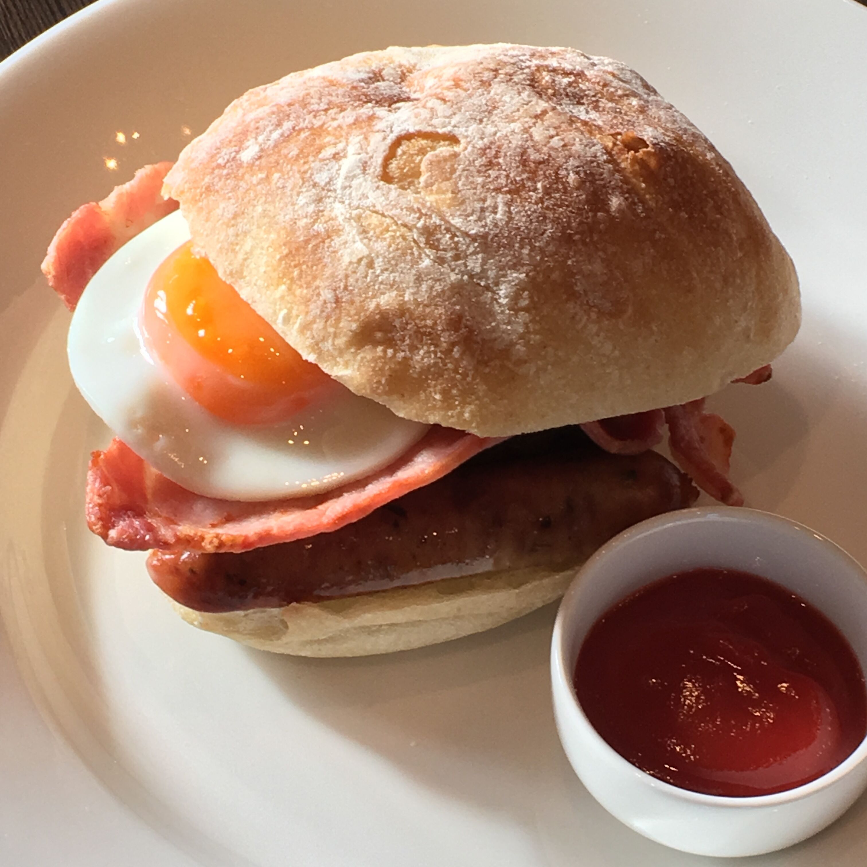 Un sandwich petit-déjeuner composé de jambon, de bacon et d’un œuf au plat, servi dans un pain et accompagné d’une portion de ketchup.