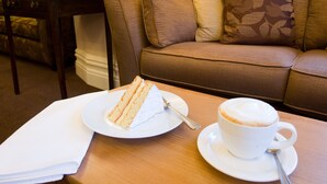 Une table à manger avec une part de gâteau et une tasse de café, un canapé marron avec des coussins à motifs et une table d’appoint en bois avec une lampe et un vase décoratif.
