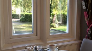 Une table de petit-déjeuner avec des assiettes remplies de nourriture, une théière et des tasses de café, placée près d’une fenêtre donnant sur des arbres et une pelouse.