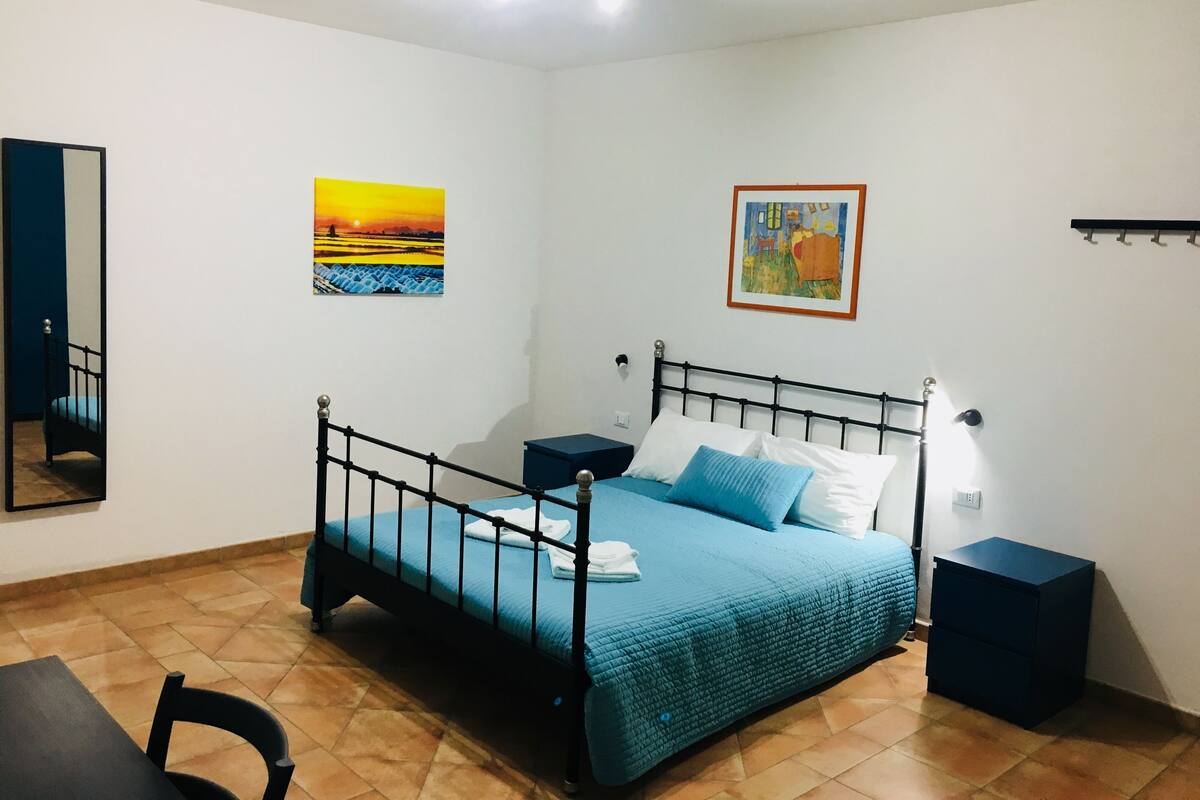 Un dormitorio con estructura de cama metálica, colcha azul, dos mesitas de noche y una lámpara de pared.
