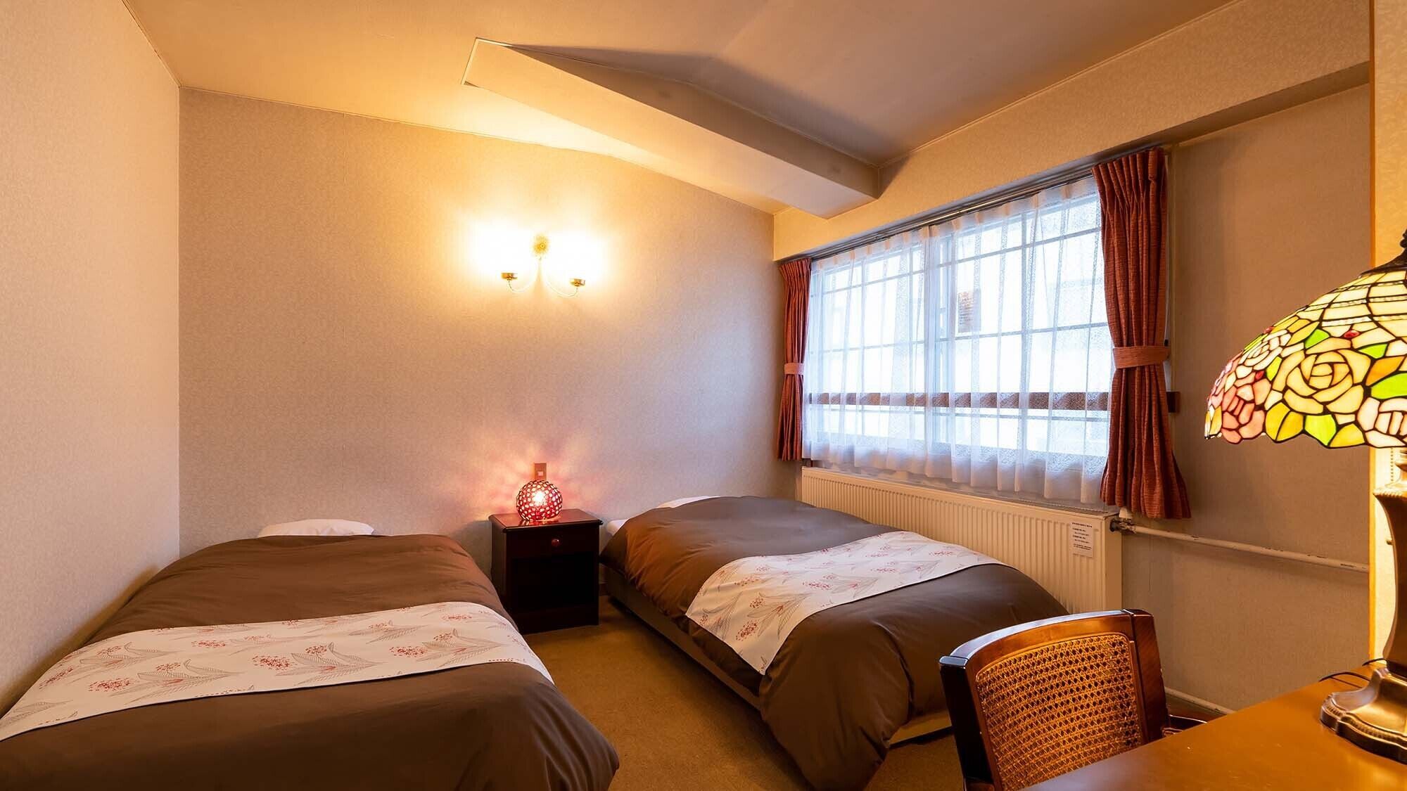 A room with two beds, a nightstand with a lamp, a chair, and a window with curtains.