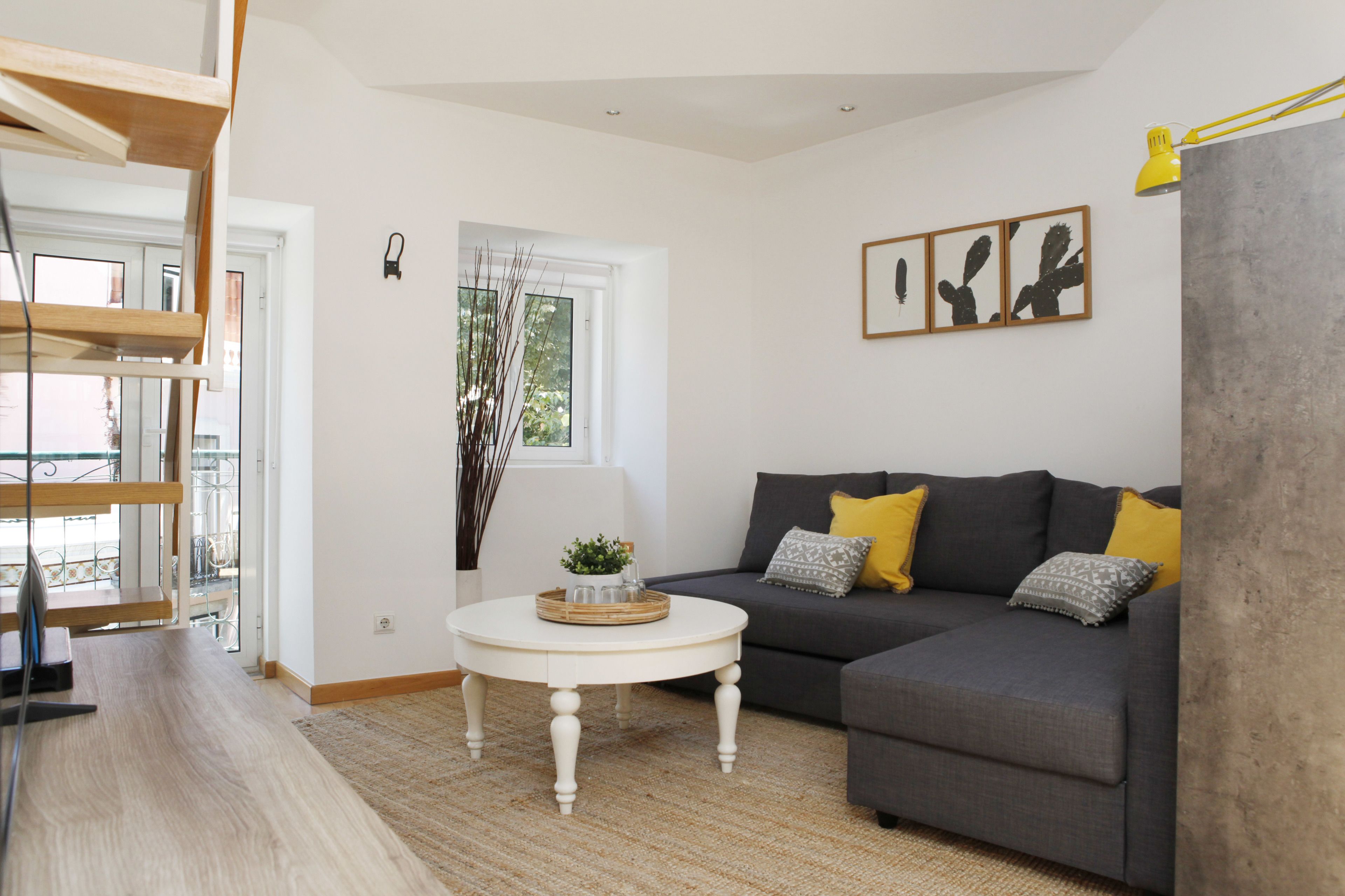 Una sala de estar moderna con un sofá gris, cojines amarillos, una mesa de centro blanca y una obra de arte con un cactus enmarcado en la pared.