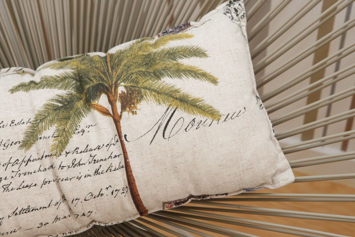 Almohada con diseño de palmera y texto escrito a mano, sobre una silla de estructura metálica.