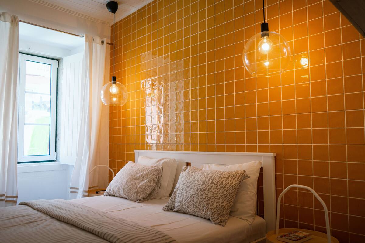 Een hotelkamer met een bed, witte gordijnen, een raam en oranje betegelde muren.