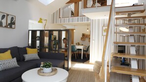 Un espace de vie moderne de style loft, avec un canapé gris foncé, des coussins jaunes, une table basse blanche agrémentée d’une plante, et un escalier menant à un niveau supérieur.