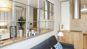 Un salon moderne avec un grand miroir, un canapé foncé et un espace cuisine en bois visible à travers le miroir.