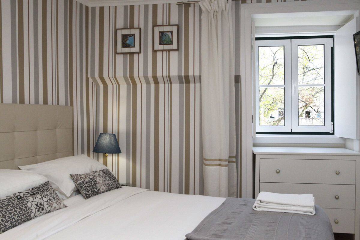 Hotelkamer met gestreepte behang, een bed met witte lakens, een bedlampje, een raam met uitzicht op bomen en een klein nachtkastje met een opgevouwen handdoek.