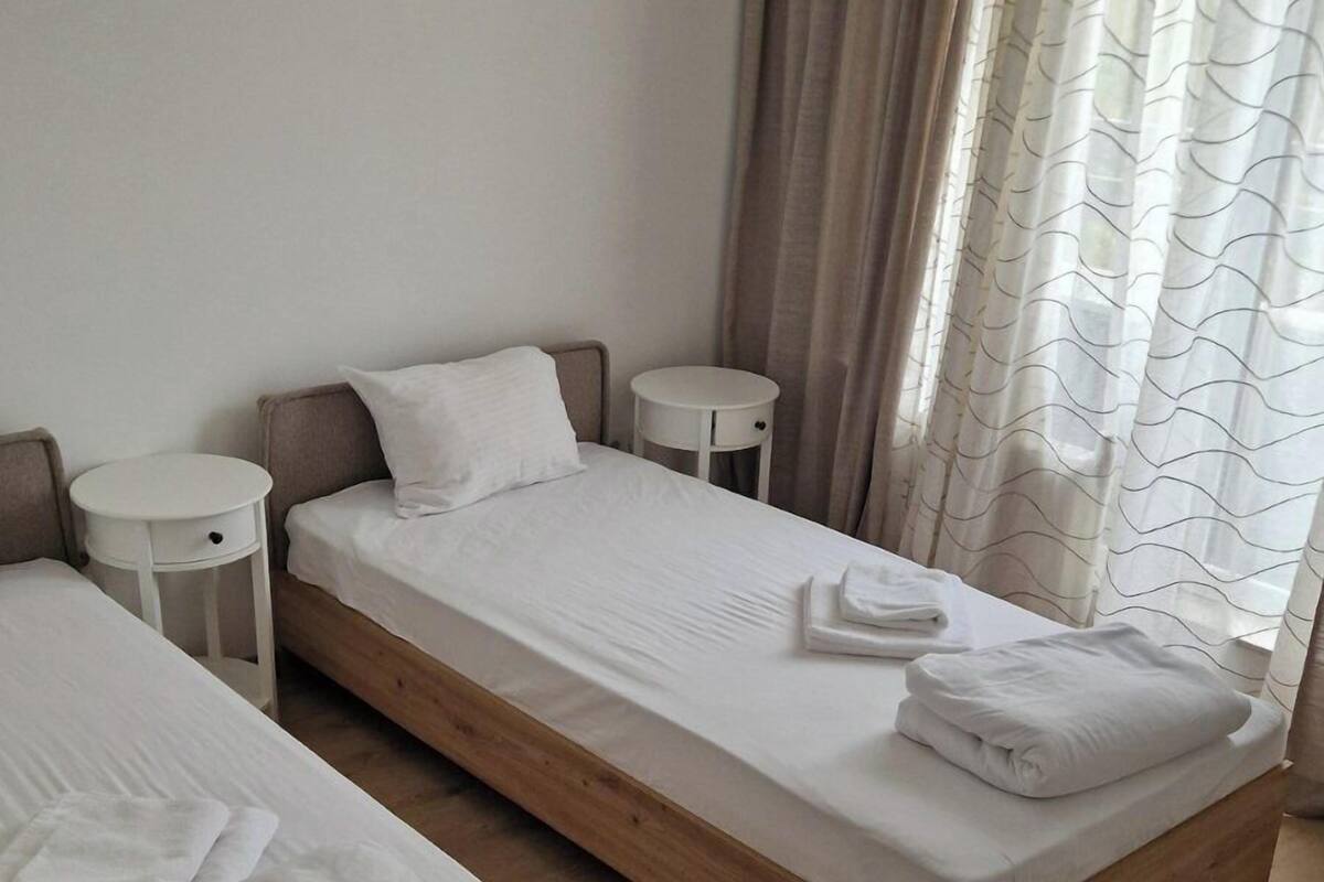 A hotel room with two single beds, a small white side table, a round white nightstand, and a window with curtains.