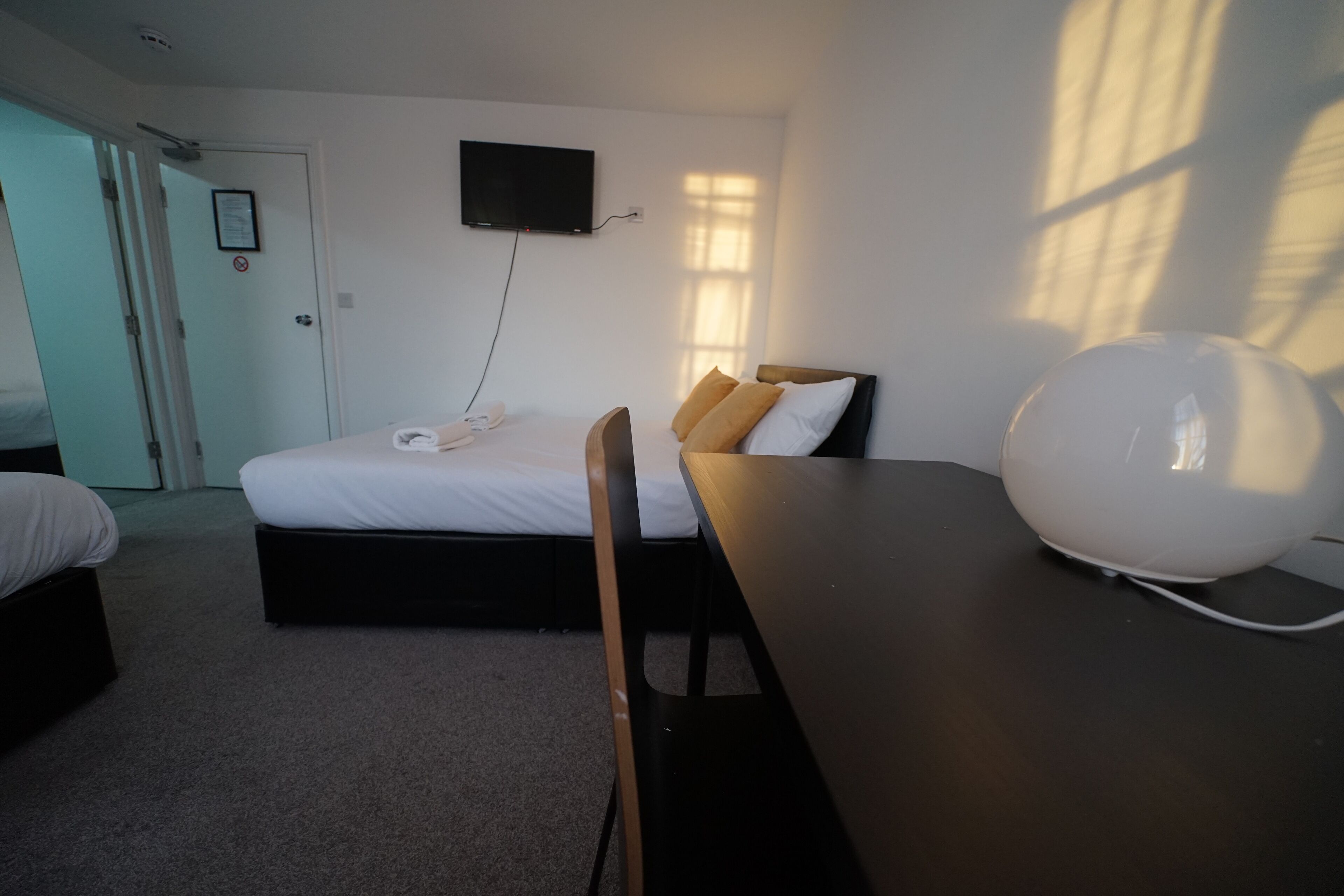 A hotel room with a bed, a desk, a chair, a flat-screen TV mounted on the wall, and a bedside table with a lamp.