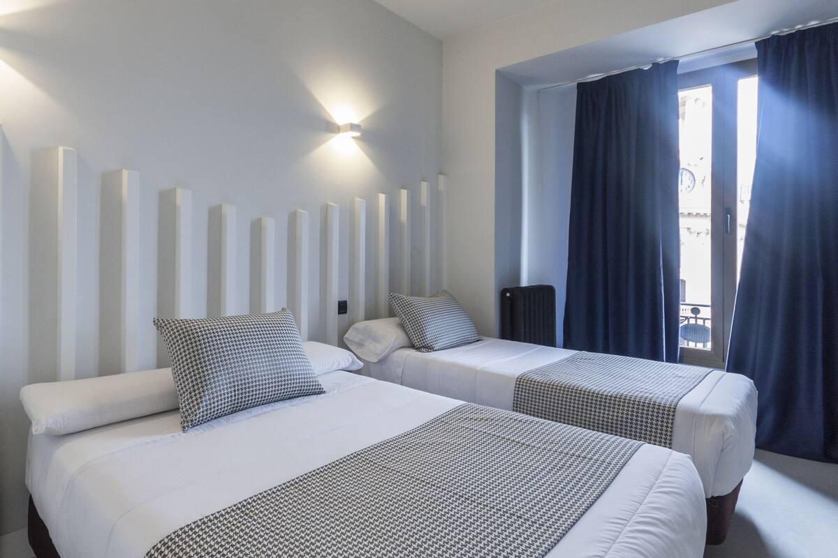 A hotel room with two beds, a wall-mounted light fixture, and a window with curtains.