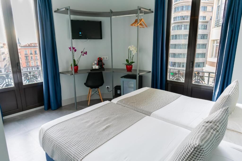 A hotel room with a bed, a desk, a chair, a TV, and a view of buildings.