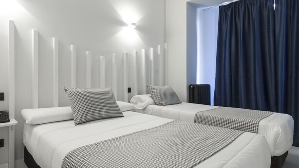 A modern hotel room with two beds, a large window with dark curtains, and a wall-mounted light fixture. - Hostal El Carmen (Madrid)
