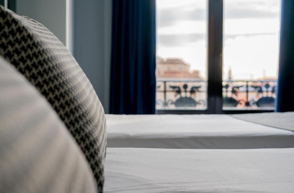 A hotel room with a bed, a window with a view of a cityscape, and a partial view of a patterned sofa.