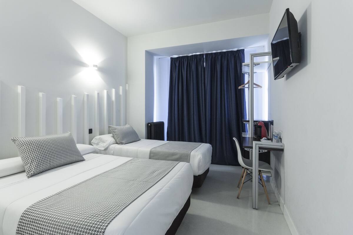 A hotel room with two beds, a desk, a chair, a flat-screen TV, and a window with curtains.
