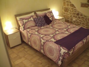 Una cama con una colcha estampada, dos almohadas y una manta. Hay dos mesitas de noche con lámparas. La habitación tiene una pared de piedra y un suelo de baldosas.