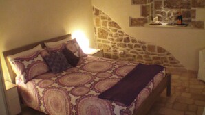 A bedroom with a stone wall, a bed with patterned bedding, a nightstand, and a small table with a bottle and glasses. - B&B La Grotta (Mesagne)