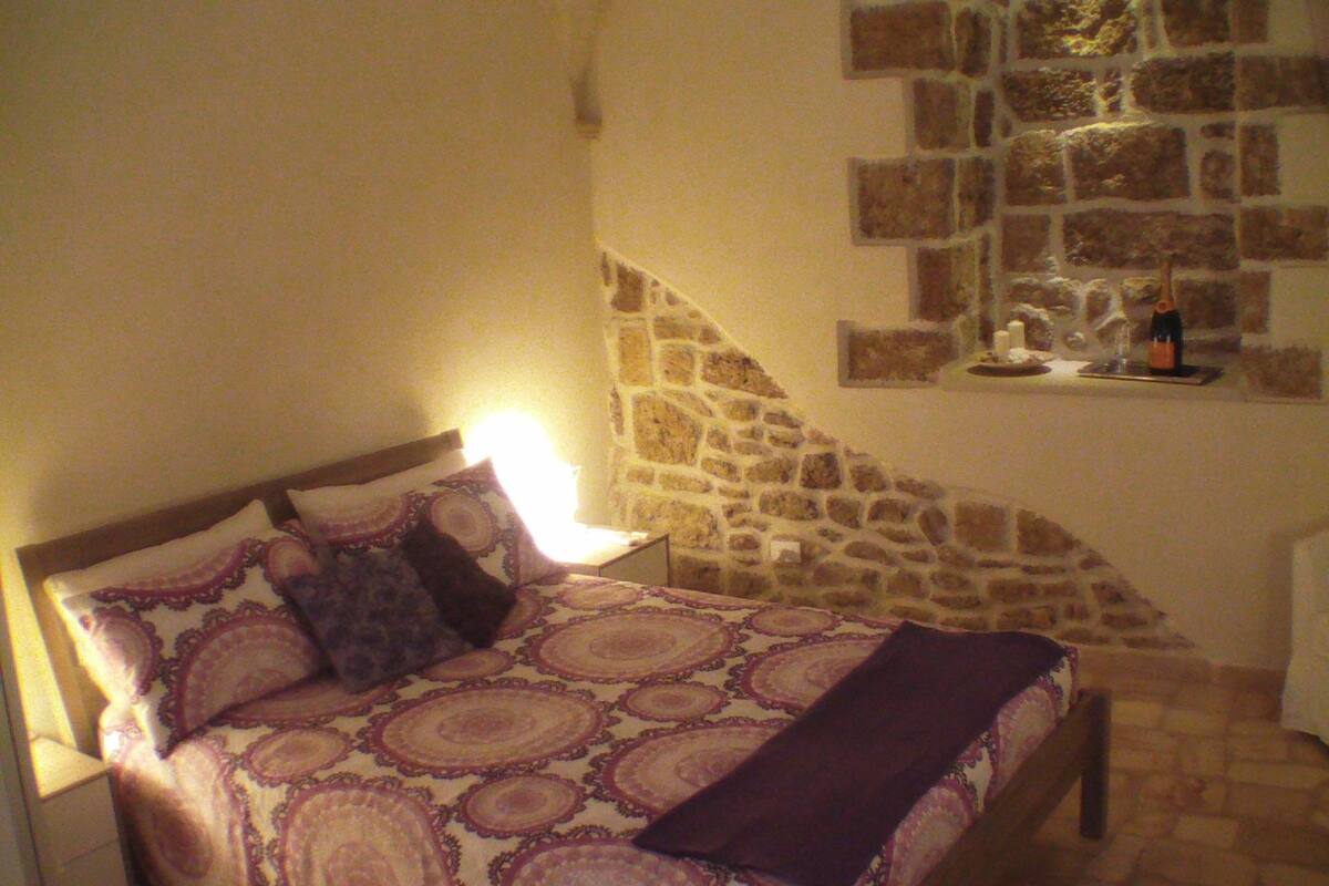 A bedroom with a stone wall, a bed with patterned bedding, a nightstand, and a small table with a bottle and glasses.