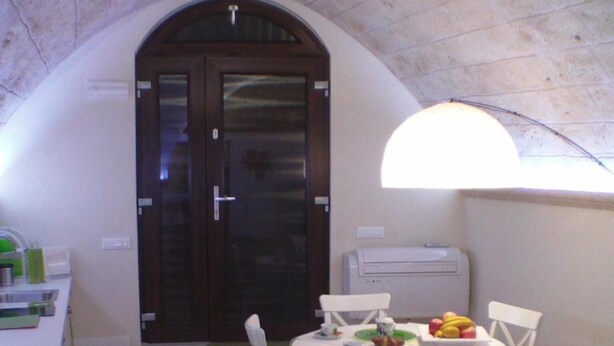 A cozy living area with a stone floor, a round table with chairs, a sofa with a green pillow, and a large arched doorway leading to a dark room. - B&B La Grotta (Mesagne)