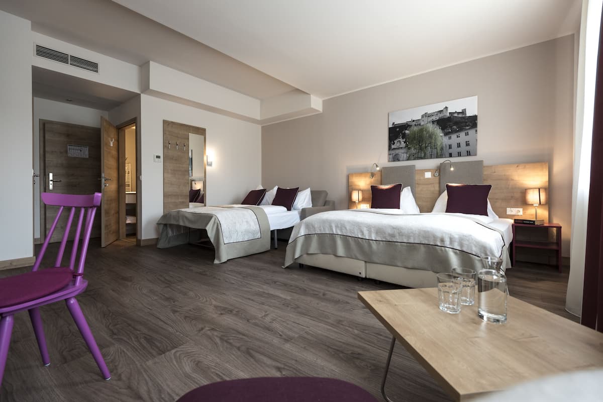 A hotel room with two beds, a wooden floor, a purple chair, and a table with glasses and water bottles.