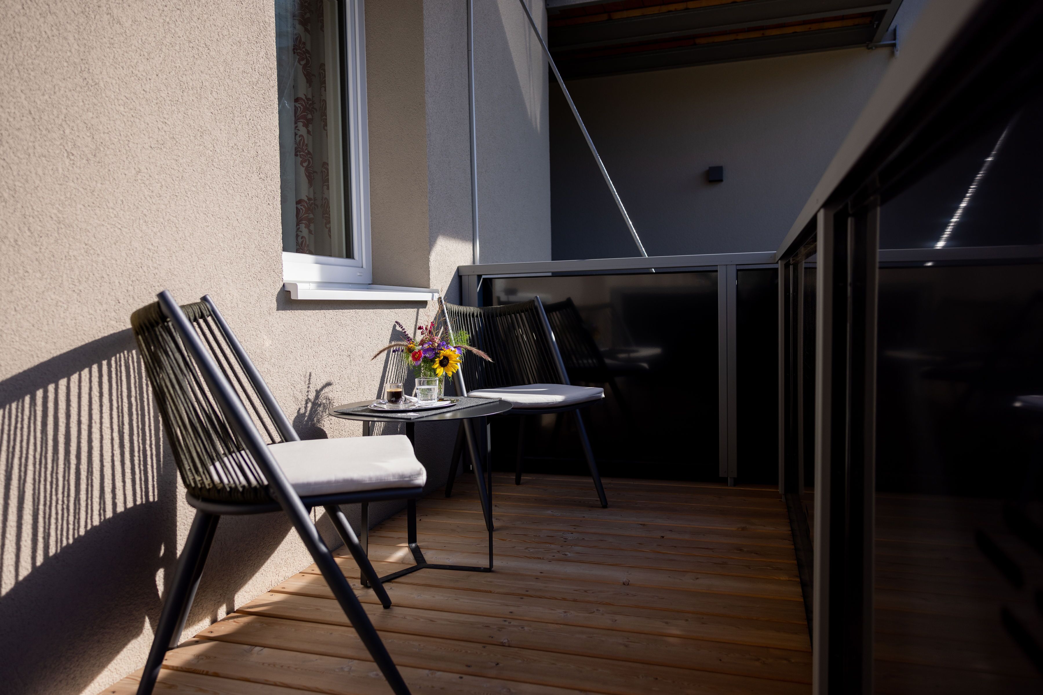 A wooden deck with two chairs and a table, a window with curtains, and a view of the building's interior.