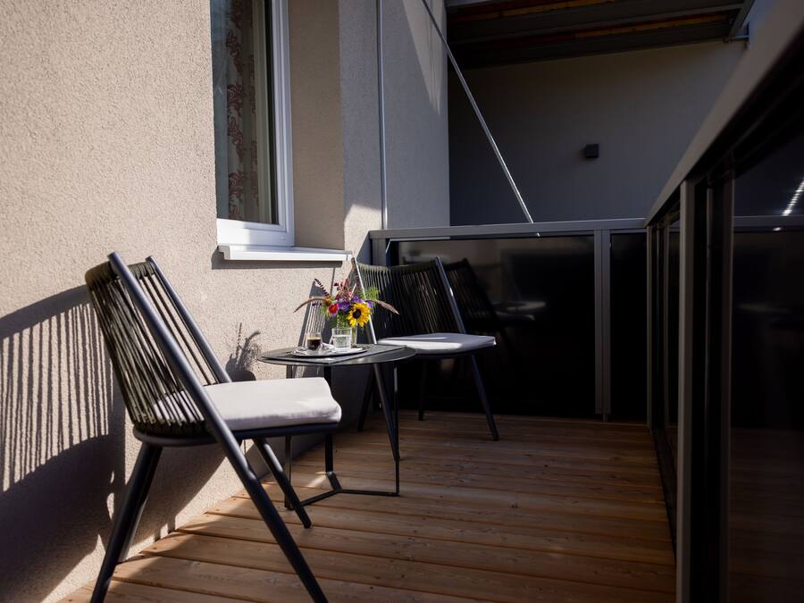 Eine Holzdauerfläche mit zwei Stühlen und einem Tisch, ein Fenster mit Vorhängen und Blick ins Innere des Gebäudes.
