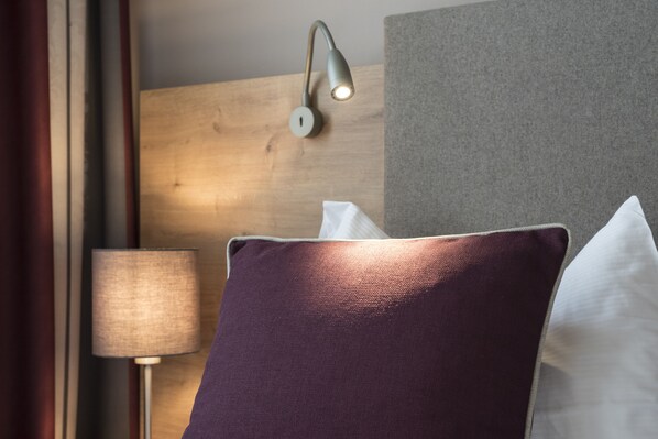 Una cama con un almohadón morado y uno blanco, una lámpara y una lámpara de pared.