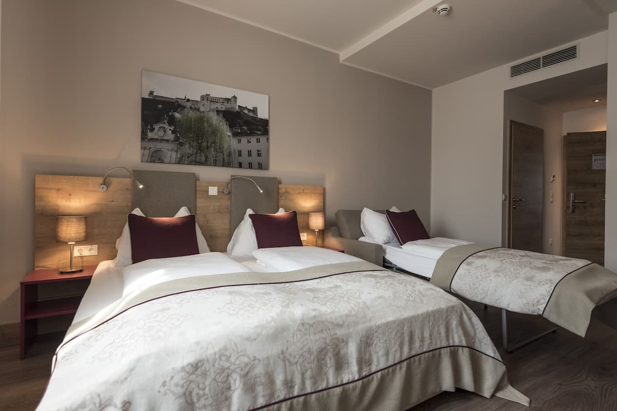 A hotel room with three beds, a wooden headboard, a wall-mounted picture, and a nightstand with a lamp.