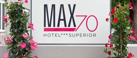 A sign for MAX 70 HOTEL SUPERIOR with a modern design and pink flowers on either side.