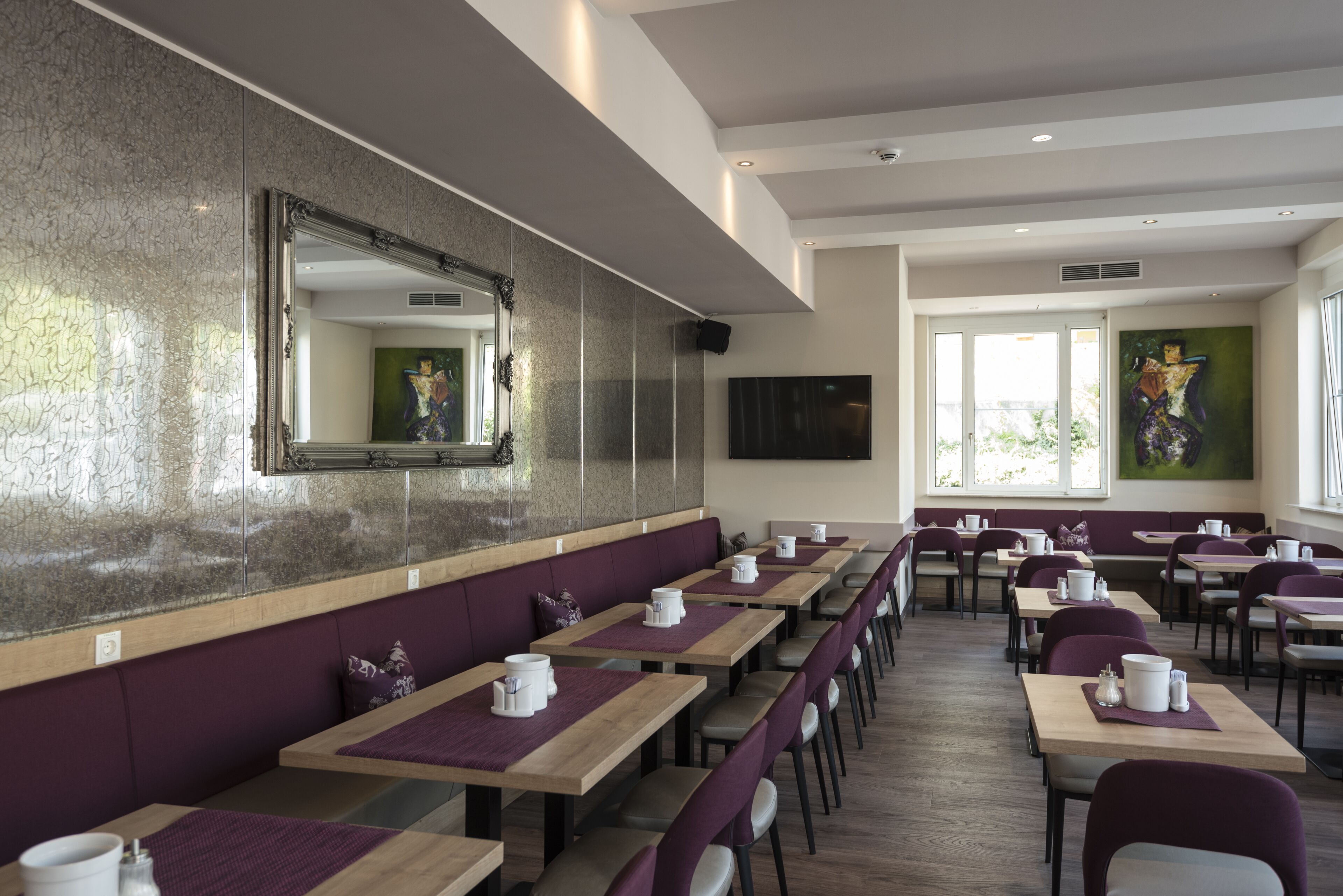 A modern restaurant with long wooden tables, purple cushioned booths, and a large mirror on the wall.