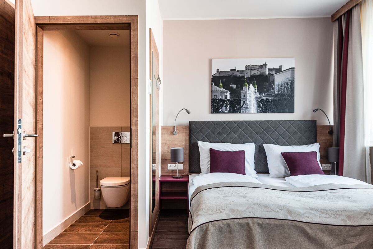 A hotel room with a bed, a nightstand, a bedside table, a picture on the wall, and a bathroom with a toilet and sink.
