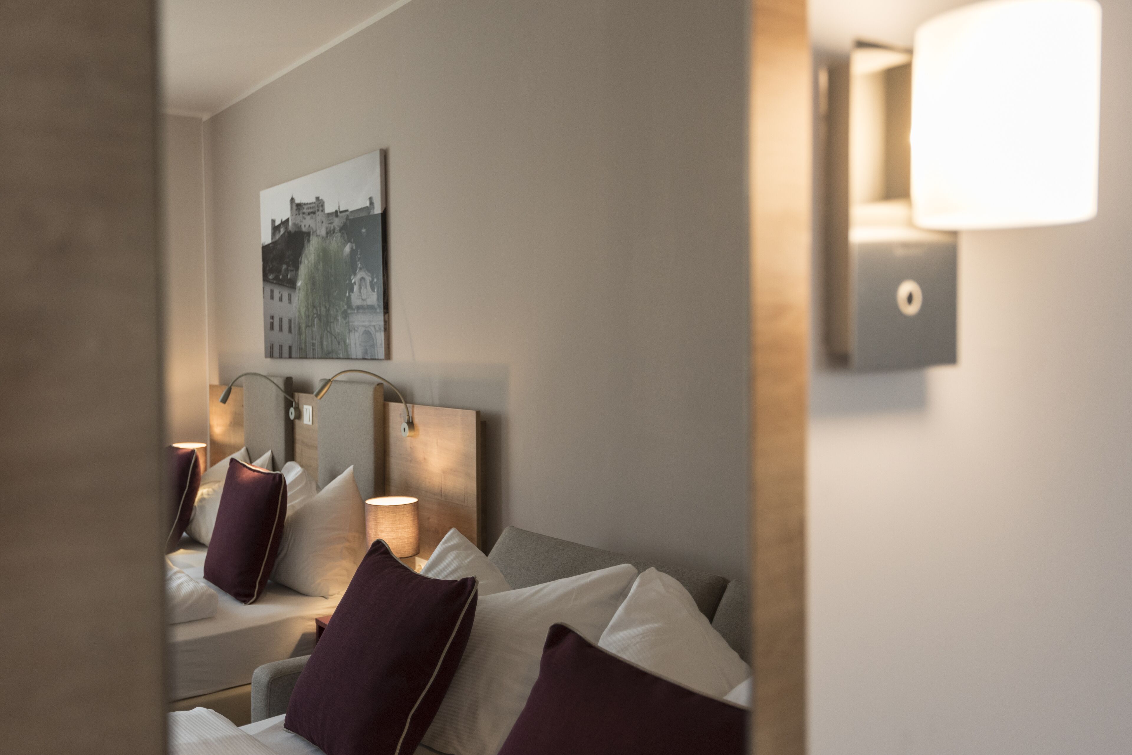 Une chambre d’hôtel avec un lit, des oreillers, une tête de lit, une lampe fixée au mur et un tableau encadré accroché au mur.