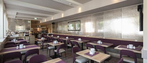 A modern dining area with multiple tables and chairs, a kitchen in the background, and a large window providing natural light.