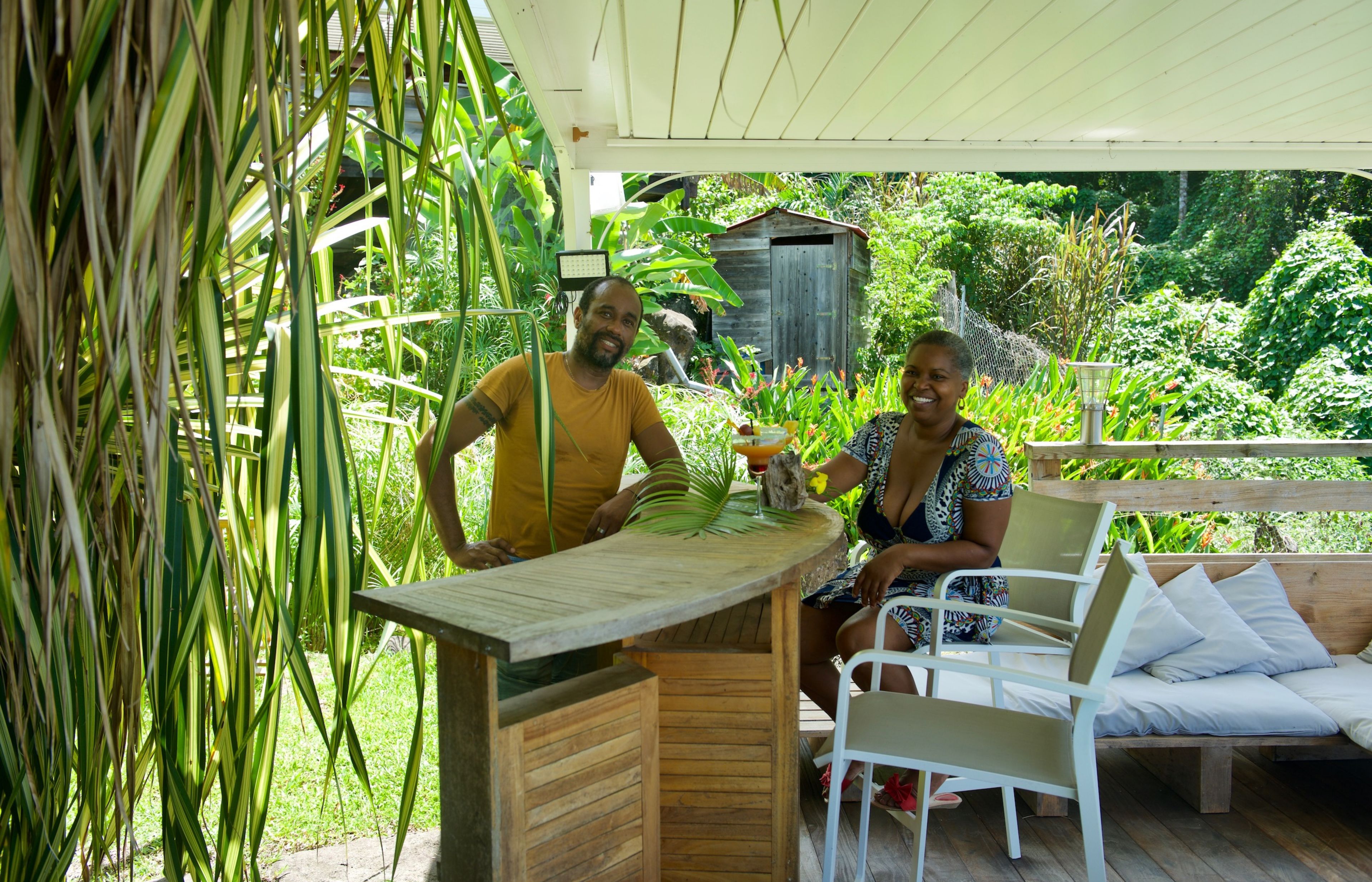A wooden outdoor seating area with a table and chairs, surrounded by lush greenery and a wooden structure in the background.