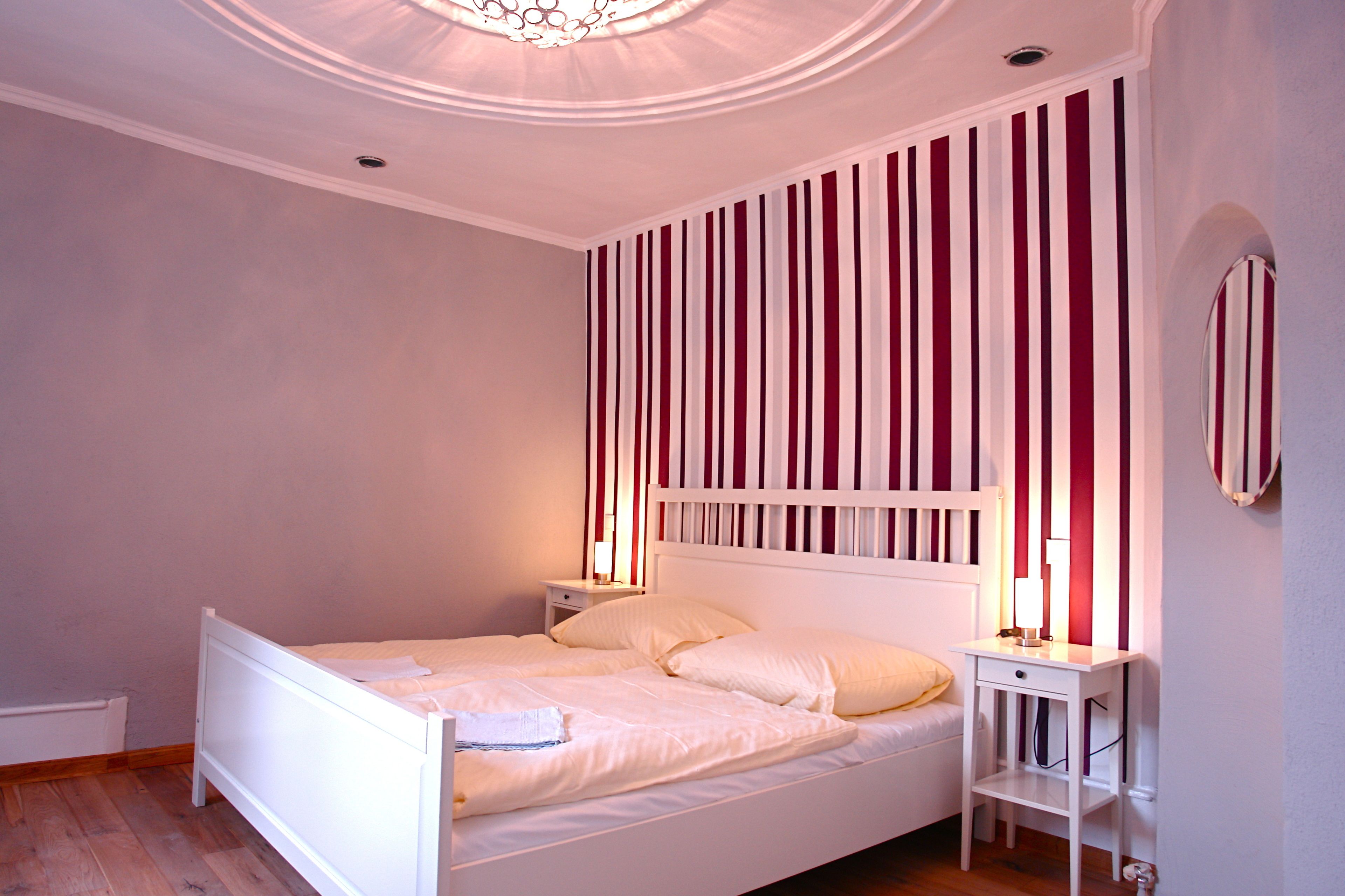 Ein Schlafzimmer mit einem weißen Bett, einem Nachttisch, einer Wandlampe und einem gestreiften Wanddesign.