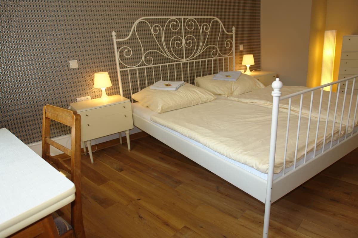 Un dormitorio con estructura metálica para la cama, dos mesitas de noche con lámparas, una silla de madera y un pequeño escritorio con silla.
