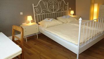 Quarto com estrutura de cama de metal, duas mesas de cabeceira com abajures, uma cadeira de madeira e uma pequena escrivaninha com cadeira.