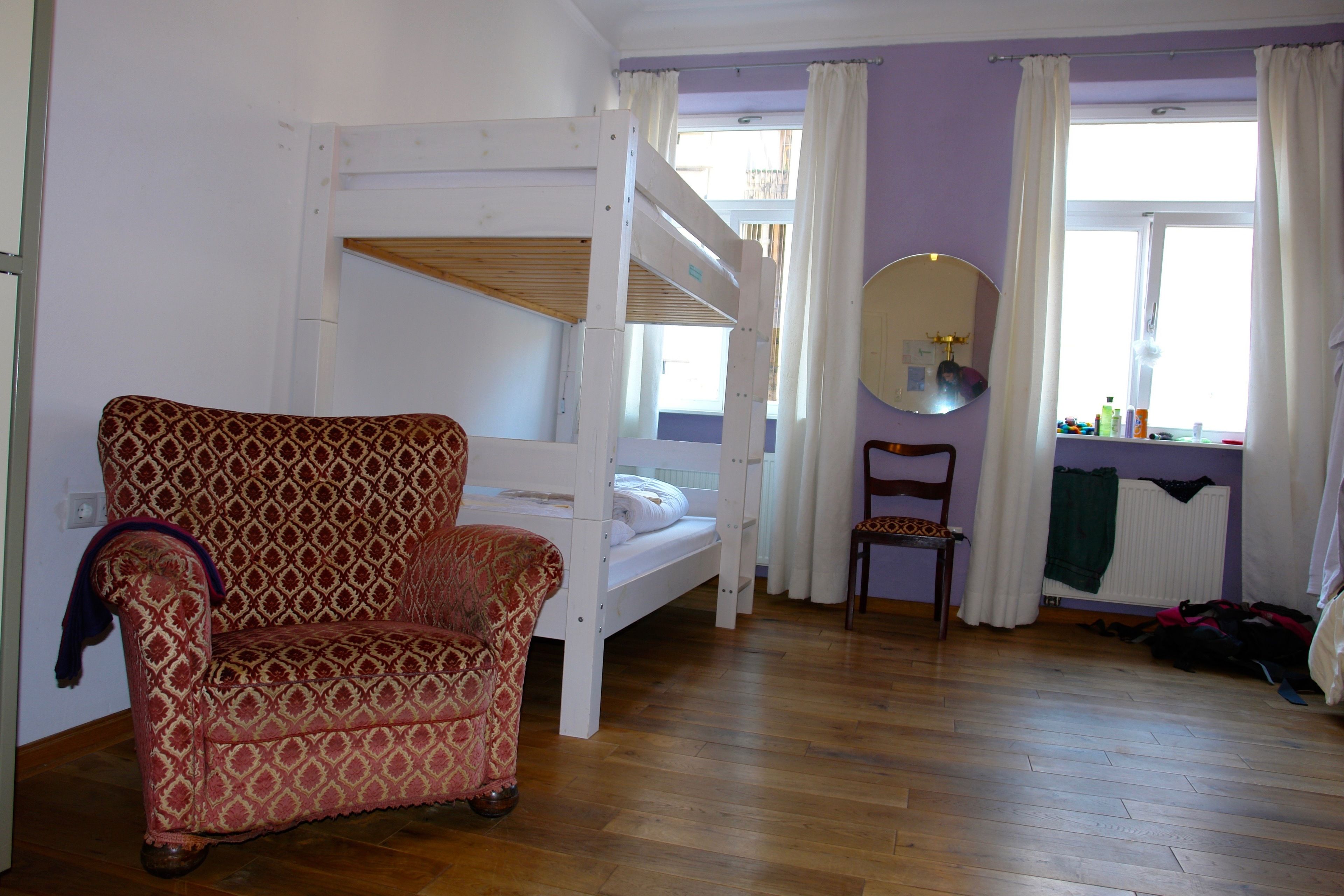 Una habitación con una cama litera, un sillón estampado, una silla, una ventana con cortinas y un espejo.