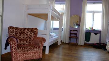 Una habitación con una cama litera, un sillón estampado, una silla, una ventana con cortinas y un espejo.