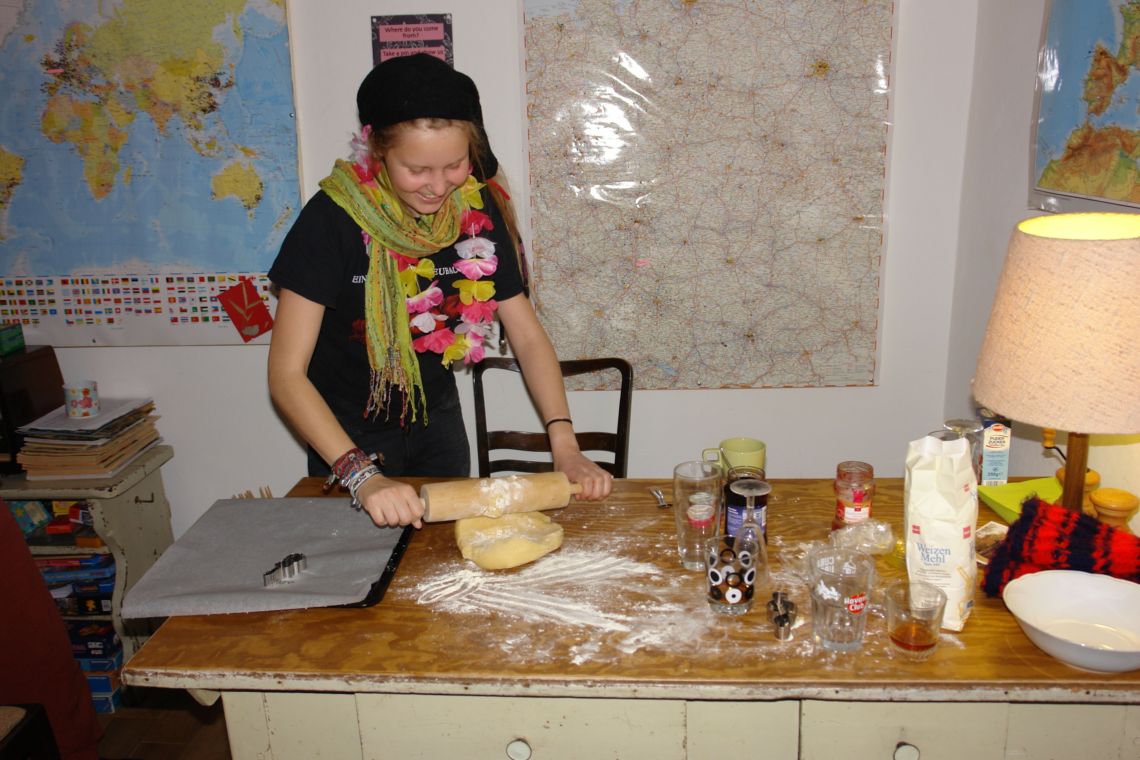 Una persona extiende masa sobre una mesa, con harina esparcida alrededor, un mapa en la pared y varios utensilios de cocina.