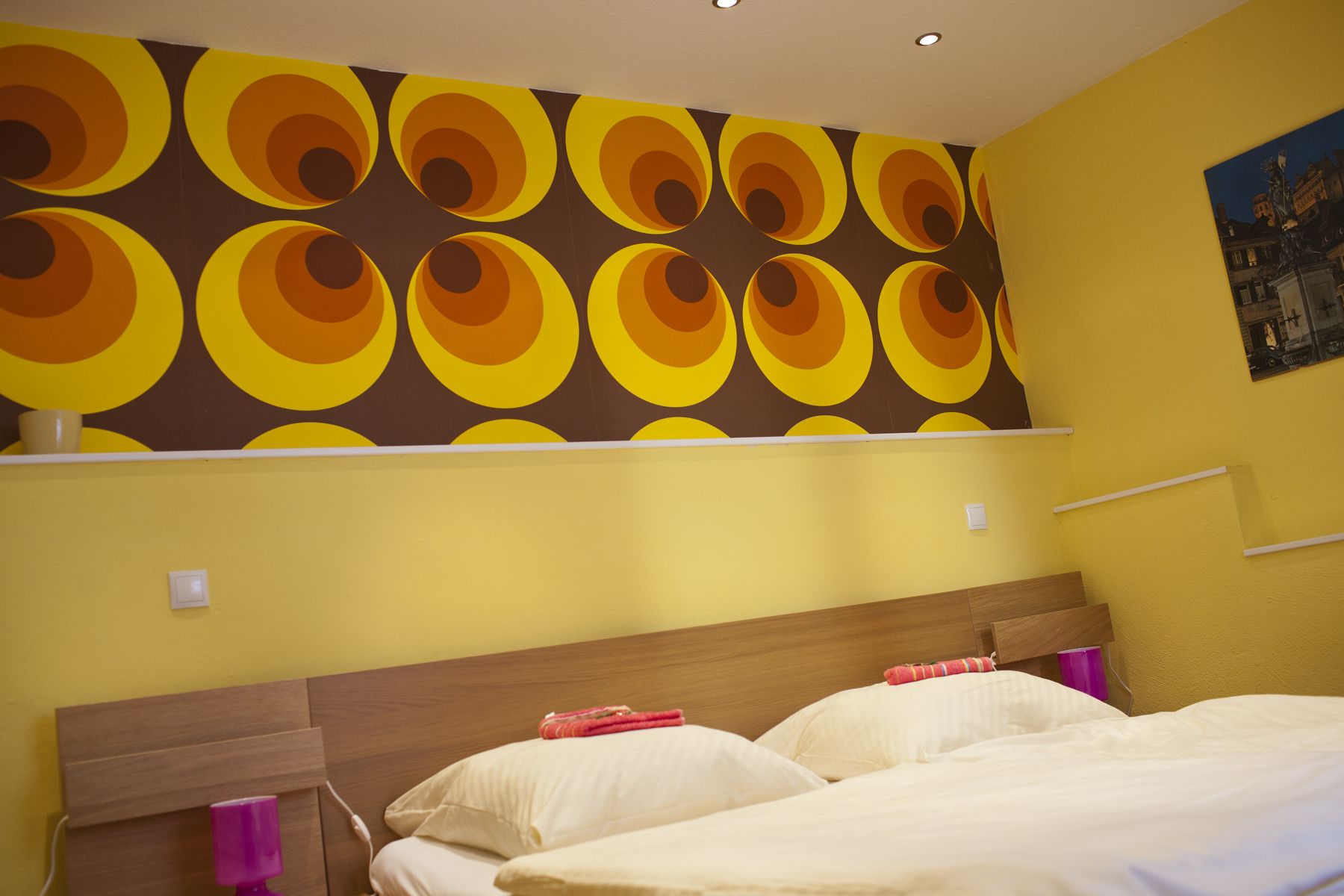 A hotel room with a bed, a wooden headboard, a wall with a circular pattern, and a painting on the wall.