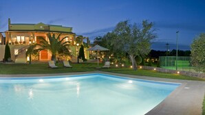 Una piscina amplia y bien iluminada con sillones de descanso y sombrillas, rodeada de un jardín ajardinado y una casa grande de varios niveles e iluminación exterior.
