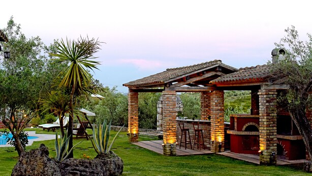Un patio con una barbacoa de ladrillo y asientos, rodeado de vegetación y una piscina.