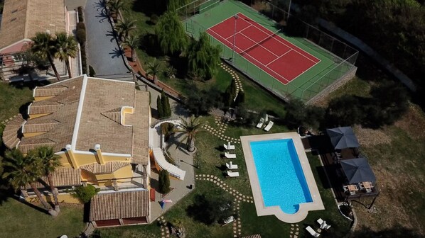 Vista aérea de una propiedad con piscina, cancha de tenis y un edificio con techo de tejas.